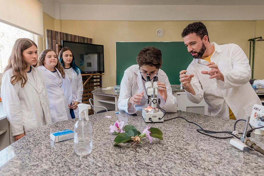 Laboratório de ciências da unidade Múltiplo, com professor e alunas de avental em experimento